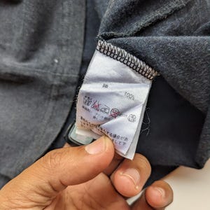 Op de afbeelding: Close-up van een donkergrijs kledinglabel met Japanse tekst en verzorgingssymbolen. Het label geeft "100%" aan en bevat wasinstructies. Het kledingstuk lijkt een t-shirt of een soortgelijk kledingstuk te zijn.