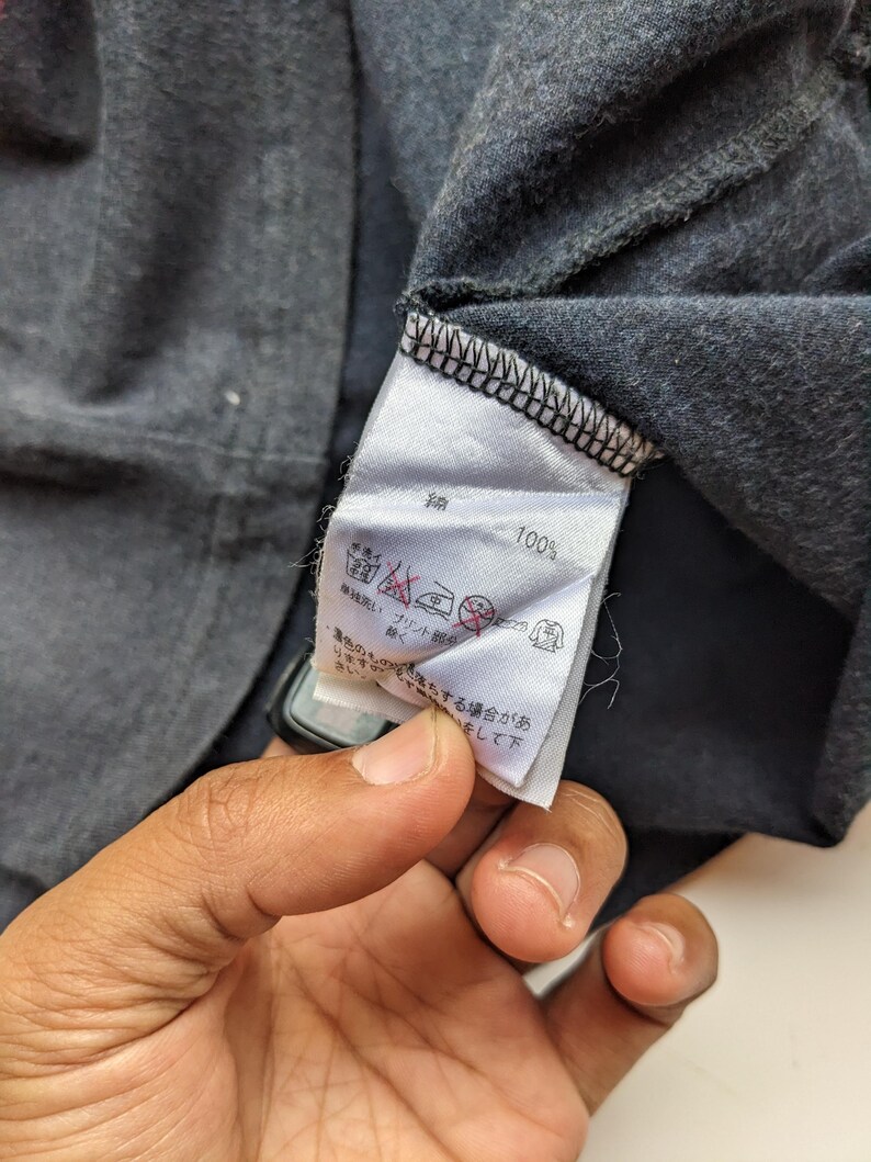 Op de afbeelding: Close-up van een donkergrijs kledinglabel met Japanse tekst en verzorgingssymbolen. Het label geeft "100%" aan en bevat wasinstructies. Het kledingstuk lijkt een t-shirt of een soortgelijk kledingstuk te zijn.