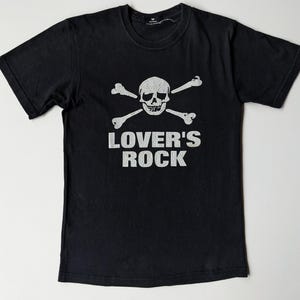 May include: Black t-shirt featuring a white skull and crossbones graphic with the text "LOVER'S ROCK". The design is centered on the front of the shirt. The shirt has short sleeves and a crew neck.