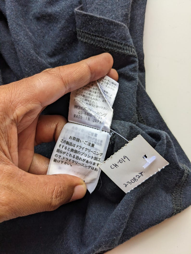 Op de afbeelding: Close-up van een donkergrijs kledingstuk met aangehechte labels. E&eacute;n label toont Japanse tekst en de woorden "MADE IN JAPAN". Een ander label heeft de tekst "CH-019" en "230825."