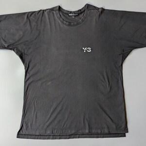 以下が含まれることがあります： 胸に白い「Y-3」ロゴが入った、ダークグレーの半袖Tシャツ。クルーネックでリラックスフィット。生地はウォッシュ加工が施されており、ヴィンテージ感があります。着回しのきくアイテムです。