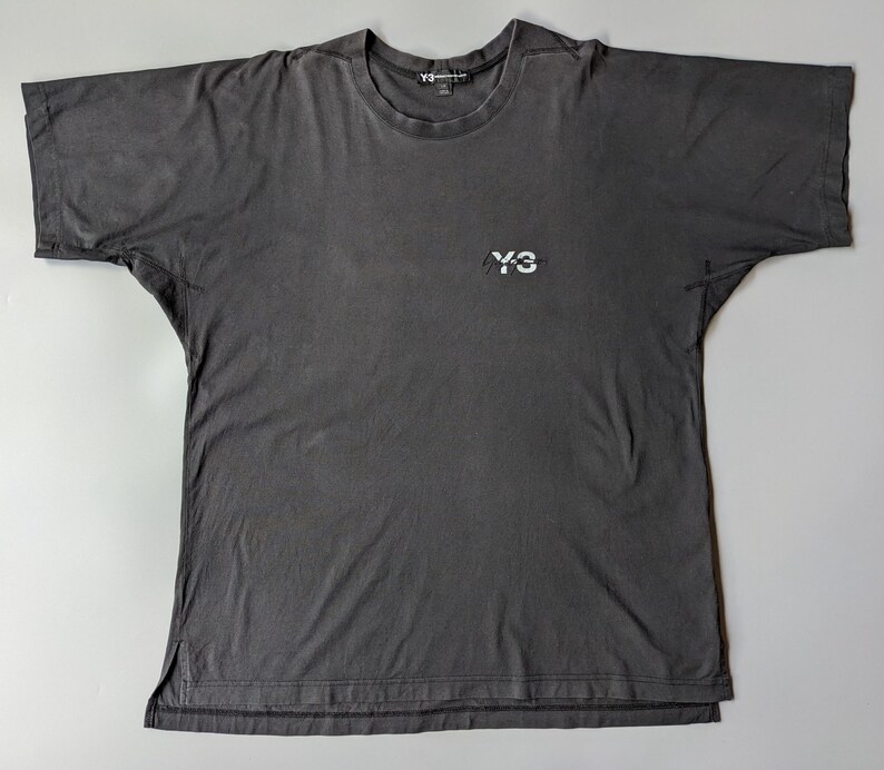 以下が含まれることがあります： 胸に白い「Y-3」ロゴが入った、ダークグレーの半袖Tシャツ。クルーネックでリラックスフィット。生地はウォッシュ加工が施されており、ヴィンテージ感があります。着回しのきくアイテムです。