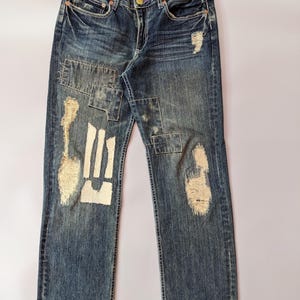 Puede incluir: Vaqueros de mezclilla azules con detalles desgastados y parches. Los vaqueros tienen un lavado descolorido, desgaste visible y múltiples parches de varias formas y tamaños. Los parches son de un tono más claro de azul y beige. Los vaqueros tienen un diseño clásico de cinco bolsillos.