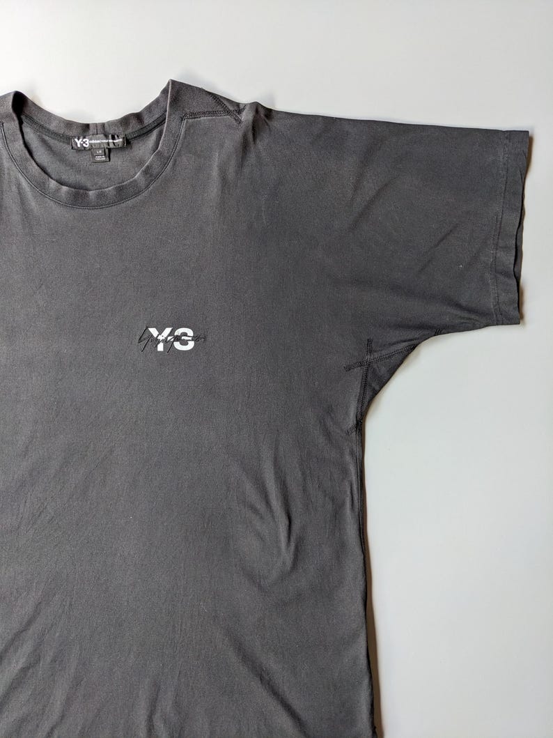 以下が含まれることがあります： クルーネックと半袖のチャコールグレーのTシャツ。胸元には白い「Y-3」のロゴが入っています。Y-3のロゴは、首の後ろのタグにも表示されています。柔らかい素材で作られたTシャツです。