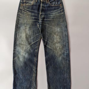 May include: Faded blue denim jeans with a button fly and a classic five-pocket design. The jeans show signs of wear, with faded areas and subtle distressing. The straight-leg cut and worn appearance give them a vintage aesthetic. The jeans are made of denim.