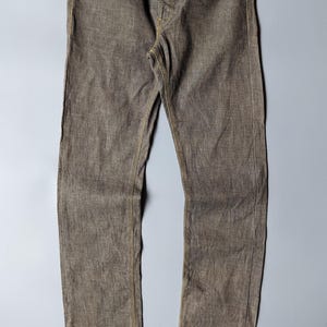 May include: A pair of brown denim jeans featuring a straight-leg cut, a button fly, and contrasting yellow stitching throughout. The waistband includes a black label with the white text RAF. The fabric has a textured, woven appearance.
