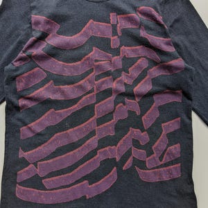 Op de afbeelding: Donkergrijs shirt met lange mouwen, een ronde hals en een groot, abstract ontwerp in paarse tinten. Het ontwerp bestaat uit overlappende, gebogen lijnen. Het shirt is gemaakt van een zacht, comfortabel materiaal.