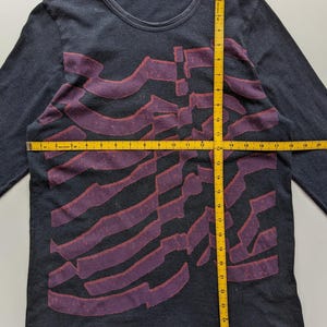Op de afbeelding: Donkergrijs shirt met lange mouwen, een ronde hals en een paars abstract ontwerp over de voorkant. Het shirt heeft de tekst "for living" op het label. Het shirt wordt gemeten met een gele meetlint.