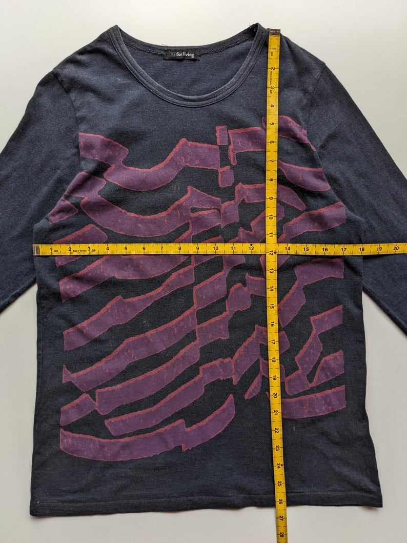 Op de afbeelding: Donkergrijs shirt met lange mouwen, een ronde hals en een paars abstract ontwerp over de voorkant. Het shirt heeft de tekst "for living" op het label. Het shirt wordt gemeten met een gele meetlint.