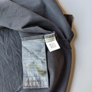 以下が含まれることがあります： ダークグレーのTシャツのクローズアップ。タグが付いており、日本語を含む複数の言語でテキストが記載されています。洗濯表示の記号も表示されています。Tシャツは柔らかく、おそらく合成素材で作られています。