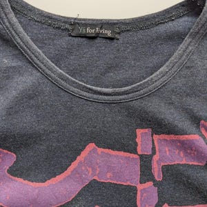 Op de afbeelding: Donkergrijs T-shirt met een ronde hals en een groot, abstract paars ontwerp met rode contouren. Het label bij de halslijn luidt "Y's for living". Het shirt lijkt gemaakt van een zacht, versleten materiaal.