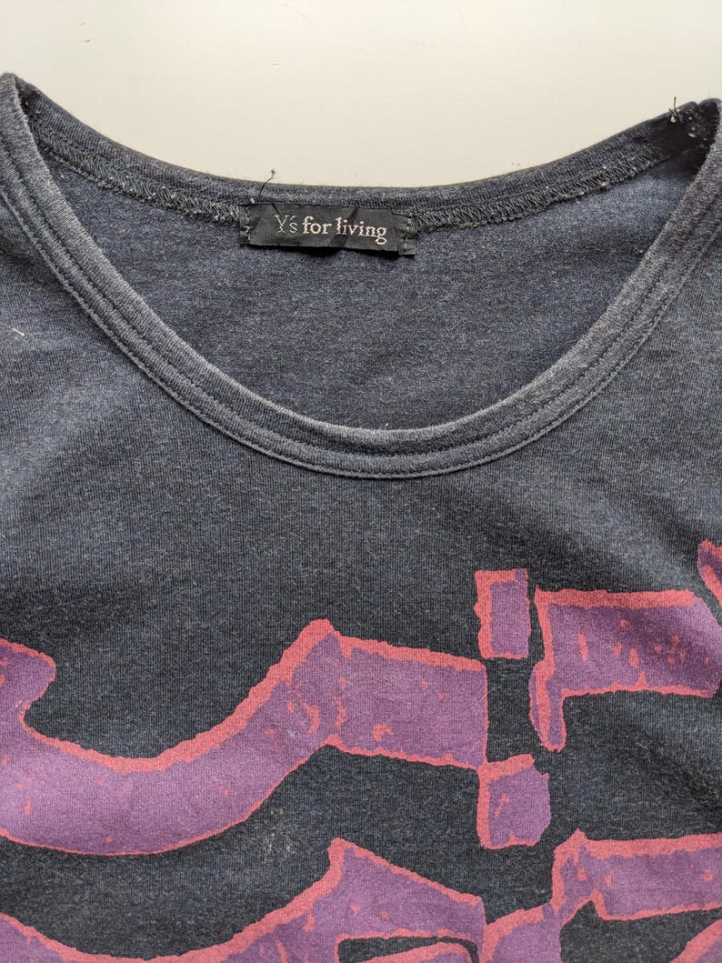 Op de afbeelding: Donkergrijs T-shirt met een ronde hals en een groot, abstract paars ontwerp met rode contouren. Het label bij de halslijn luidt "Y's for living". Het shirt lijkt gemaakt van een zacht, versleten materiaal.