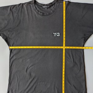 以下が含まれることがあります： クルーネックと半袖のダークグレーのTシャツ。胸元に「Y-3」のロゴ、首の後ろに「Y-3」のロゴが入ったタグが付いています。Tシャツの幅は約66cmです。