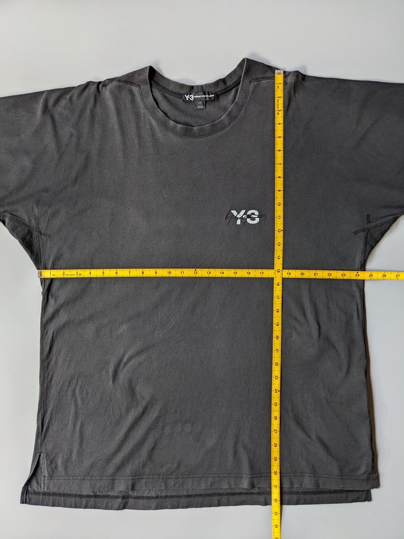 以下が含まれることがあります： クルーネックと半袖のダークグレーのTシャツ。胸元に「Y-3」のロゴ、首の後ろに「Y-3」のロゴが入ったタグが付いています。Tシャツの幅は約66cmです。