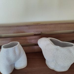 May include: Two white ceramic planters shaped like human body parts. One planter is shaped like a torso and the other is shaped like hips.