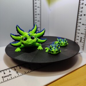 Peut inclure: Trois figurines d'axolotl vertes et bleues. Le plus grand axolotl est assis sur une base circulaire noire. Les deux plus petits axolotls sont assis sur la base derrière le plus grand axolotl.