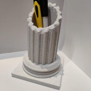 Op de afbeelding: Een witte keramische pennenhouder in de vorm van een Romeinse zuil met een vierkante basis. Twee pennen zitten in de houder, een zwarte en een gele.