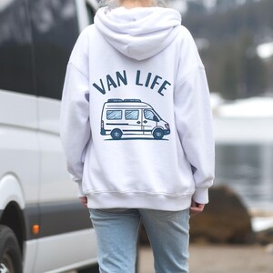 May include: White hoodie with the text "VAN LIFE" and a graphic of a camper van in blue. The hoodie is worn by a person standing in front of a white van, with a lake and mountains in the background.