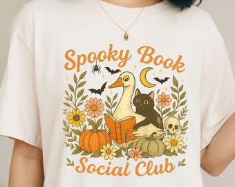 Könnte beinhalten: Cremefarbenes T-Shirt mit einem Halloween-Motiv. Das Design zeigt eine Gans, die ein Buch liest, mit einer schwarzen Katze, Kürbissen, Blumen, Fledermäusen, einer Spinne, einem Totenkopf und dem Text "Spooky Book Social Club" in Orange.