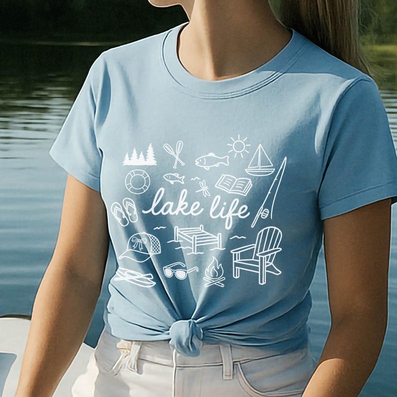 K&ouml;nnte beinhalten: Hellblaues T-Shirt mit wei&szlig;em Grafikdesign mit See-Themen-Symbolen und den Worten "lake life". Das Design umfasst ein Segelboot, einen Fisch, eine Sonne und andere Outdoor-Elemente. Das T-Shirt ist an der Taille geknotet.