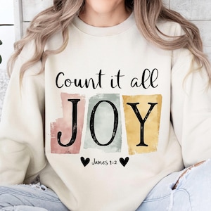 May include: Cream-colored sweatshirt with the text "Count it all JOY" in black script. The word JOY is in large block letters, each on a different colored background. Below the word JOY are two black hearts and the text "James 1:2".
