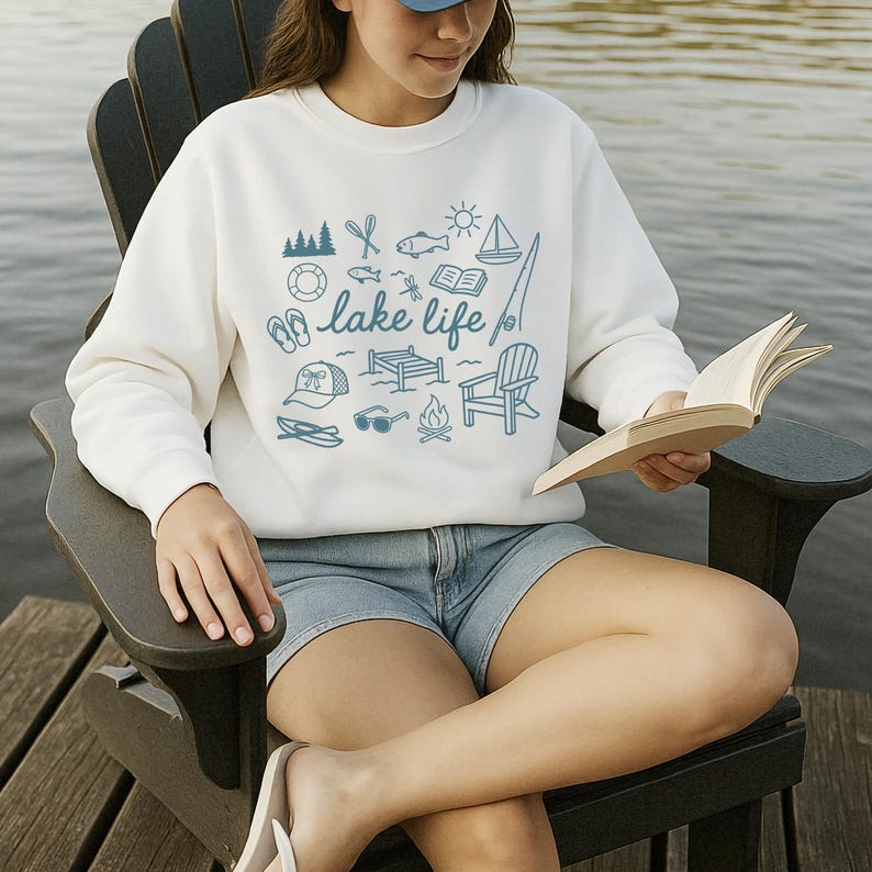 K&ouml;nnte beinhalten: Wei&szlig;es Sweatshirt mit dem Schriftzug "lake life" und blauen Illustrationen zum Thema See. Das Design umfasst ein Segelboot, eine Angelrute und ein Lagerfeuer. Die Person sitzt in einem Holzstuhl und liest ein Buch.