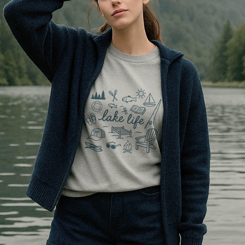 K&ouml;nnte beinhalten: Ein hellgraues Sweatshirt mit einem blauen Aufdruck "lake life" und Illustrationen zum Thema See. Die Person tr&auml;gt eine dunkelblaue Kapuzenjacke und Jeansshorts. Der Hintergrund zeigt einen See und B&auml;ume.