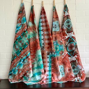 May include: Five tie-dye fabric squares in shades of brown, teal, and green. The fabric squares are hanging from clothespins on a white brick wall.