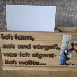 Puede incluir: Un tarjetero rectangular de madera con la frase grabada con láser Ich kam, sah und vergaß, was ich eigentlich wollte... y una pequeña ilustración en color de un pájaro a la derecha, sosteniendo una tarjeta blanca en blanco.