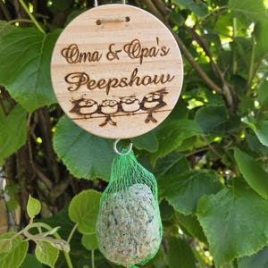 May include: A round wooden bird feeder sign with the text "Oma & Opa's Peepshow" and a row of three cartoon birds. A green mesh bag holds a birdseed cake, hanging from the sign.