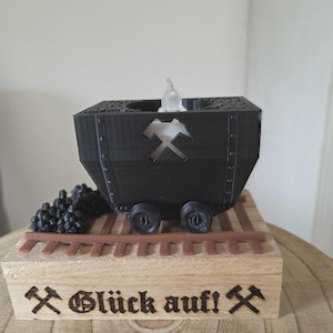 Könnte beinhalten: Ein schwarzes, 3D-gedrucktes Grubenwagen-Modell mit einer Aussparung in Form von gekreuzten Hämmern an der Seite, das auf braunen Kunststoffschienen auf einem hellen Holzsockel steht. Der Sockel ist mit dem Text "Glück auf!" und Hammer-Symbolen graviert.