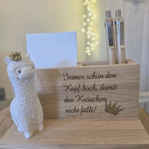 May include: A white llama figurine wearing a gold crown stands next to a wooden desk organizer. The organizer has a notepad, two pens, and the German text "Immer schön den Kopf hoch, damit das Krönchen nicht fällt!" with a crown illustration.