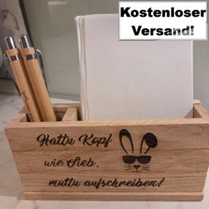 Op de afbeelding: Houten bureau-organizer met twee pennen, een stapel wit papier en een lasergeëtst ontwerp. Het ontwerp bevat een konijn met een zonnebril en de zin "Hattu Kopf wie Sieb, muttu aufschreiben!"