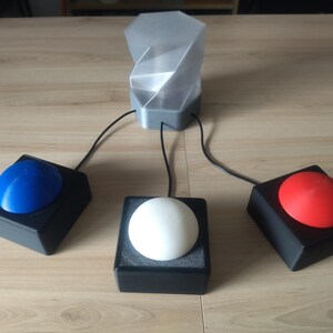 Op de afbeelding: Een futuristisch ogend apparaat met een centrale grijze structuur verbonden met drie zwarte vierkante bases. Elke basis heeft een gekleurde bol: blauw, wit en rood. Het apparaat staat op een lichtbruin houten oppervlak.