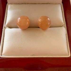 Puede incluir: Un par de pendientes de botón redondos de color melocotón expuestos en una caja de joyería de madera roja abierta con un interior de satén blanco. Los pendientes son el punto focal, sobre el elegante fondo de la caja.