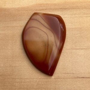 May include: A smooth, polished, brown and white agate stone with a unique, swirling pattern. The stone is shaped like a teardrop and has a glossy finish.