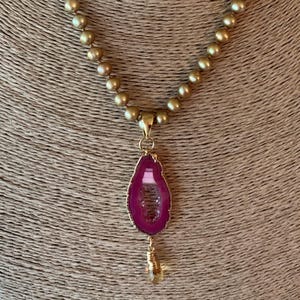 May include: A gold-toned beaded necklace with a teardrop-shaped pendant. The pendant features a vibrant pink agate stone with gold edging and a small crystal charm. The necklace is displayed on a neutral-toned mannequin.