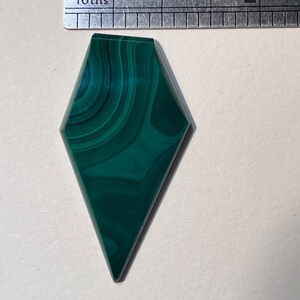 May include: A polished, diamond-shaped malachite gemstone with swirling patterns of dark and light green. A metal ruler with millimeter and sixteenth-inch markings is placed above the gemstone for scale.