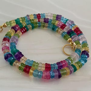 May include: A necklace composed of small, faceted, multicolored gemstone beads in shades of blue, green, pink, yellow, and purple. The strand features a gold-toned metal clasp and a small, circular gold-toned metal ring attached to the end.