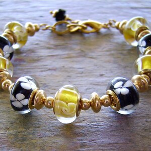 May include: A bracelet featuring alternating glass beads and gold-toned metal spacers. The glass beads include clear spheres with yellow swirls and black spheres with white floral patterns. The bracelet is secured with a gold-toned metal lobster clasp.