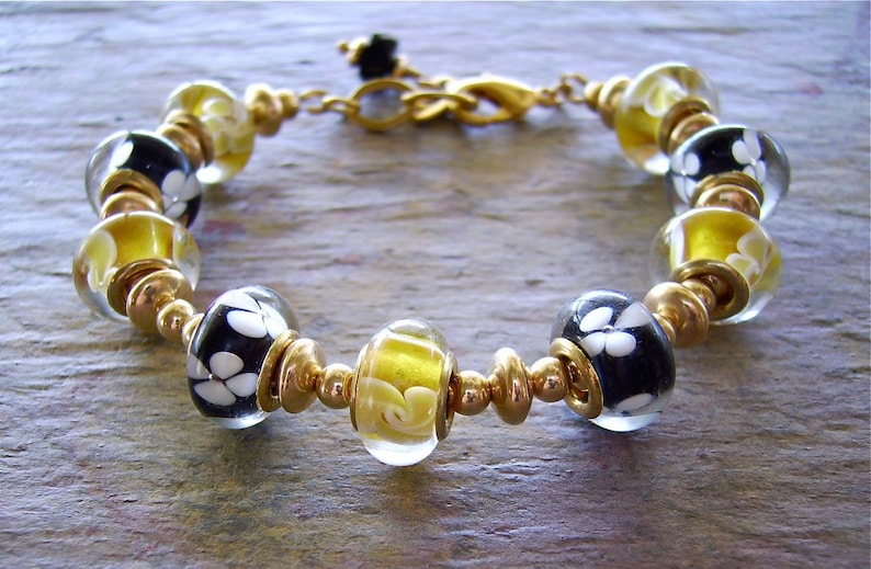 May include: A bracelet featuring alternating glass beads and gold-toned metal spacers. The glass beads include clear spheres with yellow swirls and black spheres with white floral patterns. The bracelet is secured with a gold-toned metal lobster clasp.