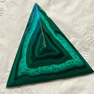 May include: A triangular malachite gemstone with concentric bands of dark and light green hues. The polished surface reflects light, showcasing the stone's natural patterns and textures. The gemstone is a vibrant display of natural artistry.