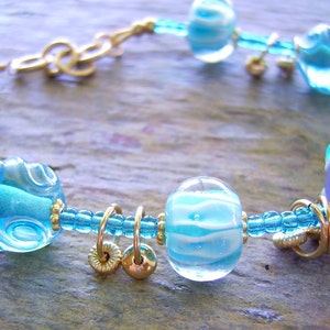 K&ouml;nnte beinhalten: Ein Armband mit durchscheinenden hellblauen und lavendelfarbenen Glasperlen mit wei&szlig;en Wirbelmustern, durchsetzt mit kleinen metallisch goldfarbenen Ringen und Perlen, verbunden durch einen d&uuml;nnen blauen Perlenstrang und einen goldfarbenen Metallverschluss.