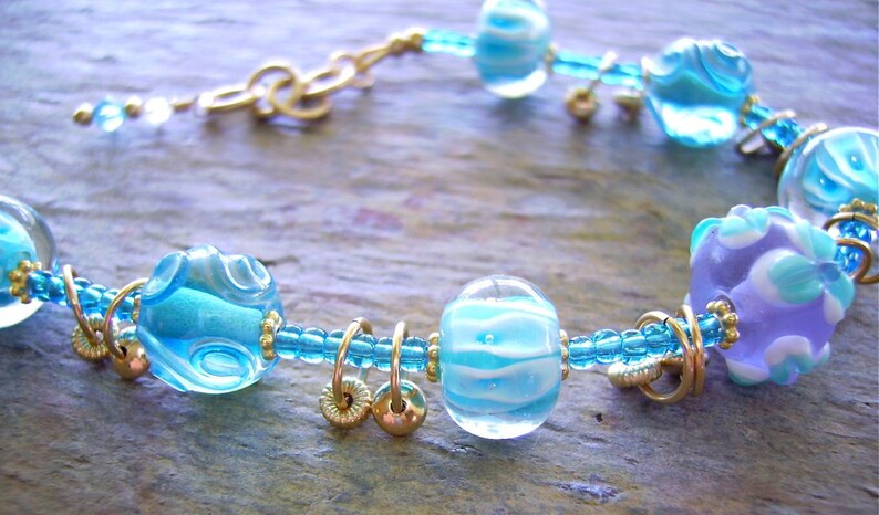 K&ouml;nnte beinhalten: Ein Armband mit durchscheinenden hellblauen und lavendelfarbenen Glasperlen mit wei&szlig;en Wirbelmustern, durchsetzt mit kleinen metallisch goldfarbenen Ringen und Perlen, verbunden durch einen d&uuml;nnen blauen Perlenstrang und einen goldfarbenen Metallverschluss.