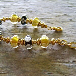 May include: A bracelet featuring alternating black and white floral patterned glass beads and translucent yellow glass beads, separated by small gold-toned metal spacer beads. The bracelet includes a gold-toned lobster clasp and a small black bead charm.