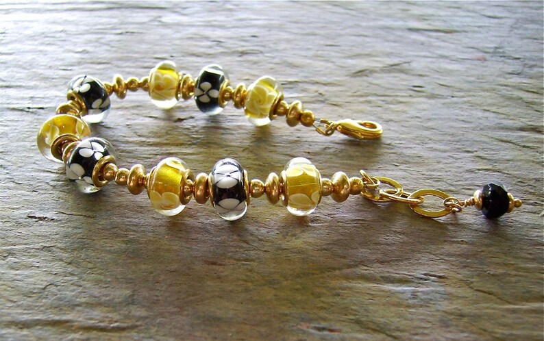 May include: A bracelet featuring alternating black and white floral patterned glass beads and translucent yellow glass beads, separated by small gold-toned metal spacer beads. The bracelet includes a gold-toned lobster clasp and a small black bead charm.