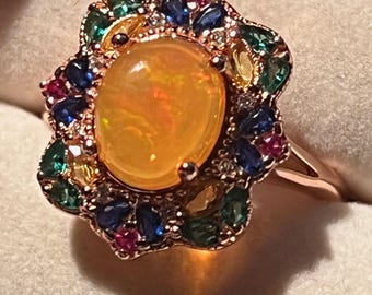 Könnte beinhalten: Ein roségoldener Ring mit einem großen, oval geschliffenen, gelben Opal. Der Opal ist von einem Kranz aus kleineren, farbigen Edelsteinen umgeben, darunter blaue, grüne und rote Steine. Der Ring hat ein poliertes Roségoldband.