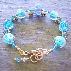 K&ouml;nnte beinhalten: Ein Armband mit hellblauen und wei&szlig;en, wirbelartig gemusterten Glasperlen, durchsetzt mit kleinen blauen Saatperlen und goldfarbenen Metallringen. Das Schmuckst&uuml;ck wird mit einem goldfarbenen Karabinerverschluss geschlossen und hat einen kleinen Anh&auml;nger.