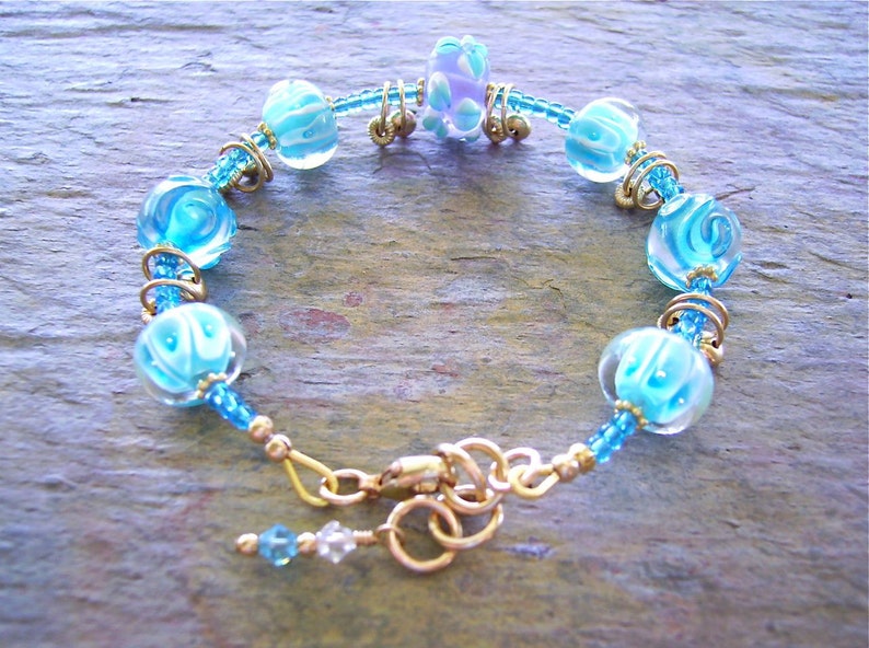 K&ouml;nnte beinhalten: Ein Armband mit hellblauen und wei&szlig;en, wirbelartig gemusterten Glasperlen, durchsetzt mit kleinen blauen Saatperlen und goldfarbenen Metallringen. Das Schmuckst&uuml;ck wird mit einem goldfarbenen Karabinerverschluss geschlossen und hat einen kleinen Anh&auml;nger.