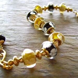 May include: A bracelet featuring alternating glass beads in black and yellow, each decorated with white floral patterns. The beads are separated by small gold-toned metal spacers and rings, finished with a gold-toned lobster clasp and a black bead.